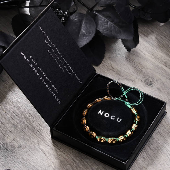 Bronze Bewitchment bracelet with bronze skulls on green and black adjustable GloWeave cord displayed in black NOGU box with care instructions